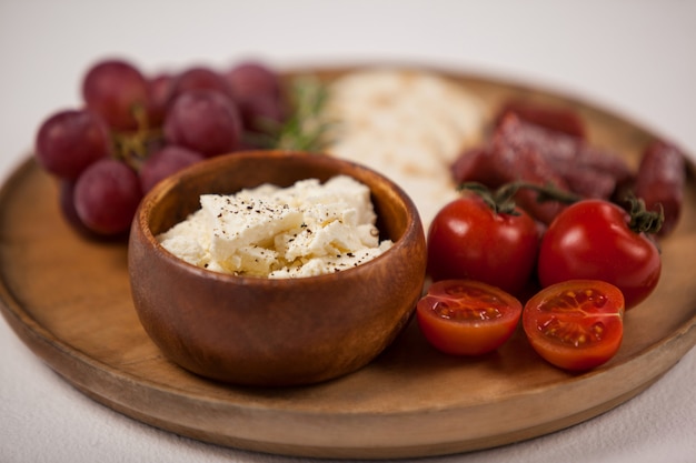 Galletas crujientes, tomates cherry, uvas y tazón de queso sobre tabla de madera