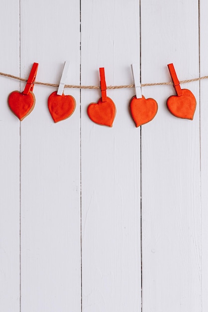 Galletas en forma de corazón con crema roja cuelgan de una cuerda sobre un fondo de madera blanca