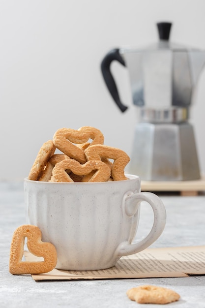 Foto galletas en forma de corazón en una taza blanca fondo claro con una cafetera