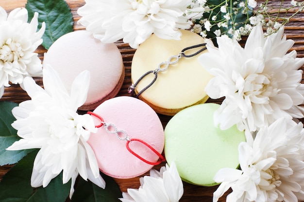 Galletas francesas de macarrón con brazaletes de mejores amigos. Bonitos accesorios con colgantes de signo de infinito presentados en dulces coloridos en marco de flor, primer plano