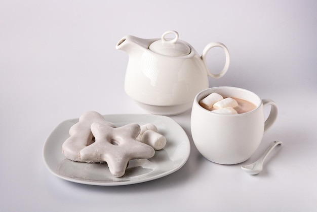 Galletas de jengibre en esmalte blanco, tetera blanca, cacao en una taza sobre un fondo blanco.