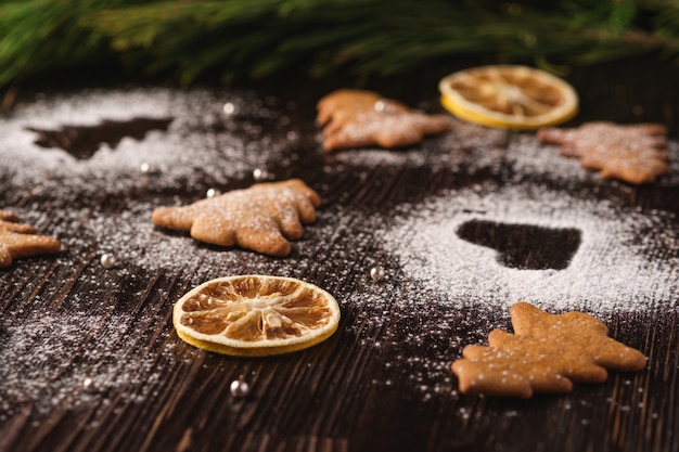 Galletas de jengibre en forma de corazón y abeto de Navidad, azúcar en polvo sobre mesa de madera, frutas secas de cítricos, rama de abeto, ángulo de visión, enfoque selectivo