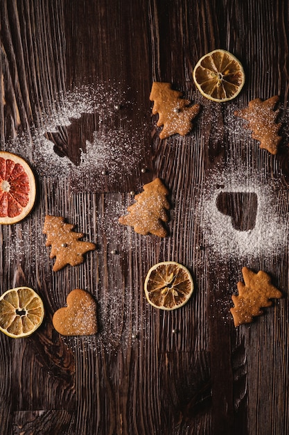 Galletas de jengibre en forma de corazón y abeto de Navidad, azúcar en polvo sobre mesa de madera, frutas secas de cítricos, vista superior