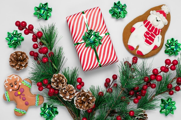 Galletas de jengibre de Navidad y adornos de año nuevo en superficie blanca. Tarjeta de felicitación con regalo, ramas de abeto, piñas y bayas de acebo rojo.