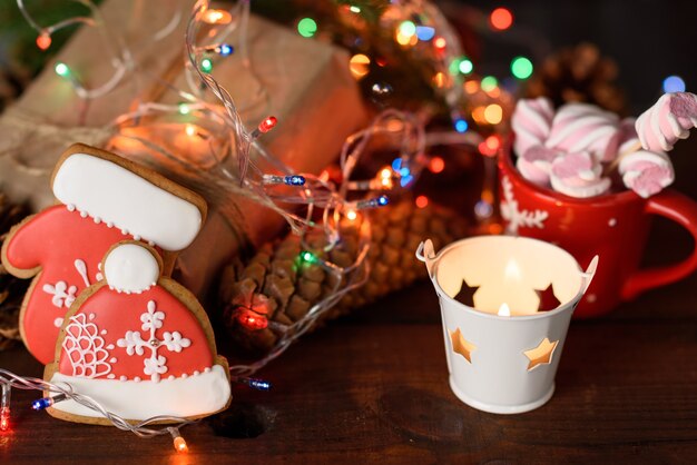 Galletas de jengibre navideñas horneadas, cacao con malvaviscos y luces navideñas encendidas sobre una mesa marrón