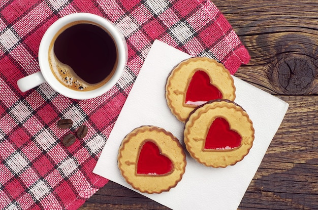 Galletas con mermelada en forma de corazones y taza de café negro sobre mantel rojo, vista superior