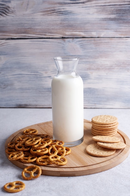 Galletas, mini pretzels salados y botella con leche sobre fondo blanco de madera.