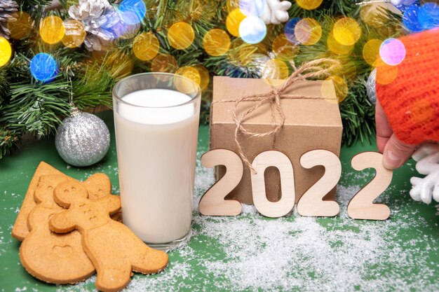 Galletas, un vaso de leche para Santa sobre una mesa cubierta de nieve en el fondo de adornos navideños y regalos. Mano que sostiene el número 2 de madera en fecha 2022. Año nuevo 2022. Fondo de Navidad.