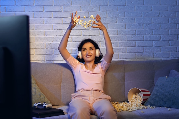 Foto gamer envolvida usando fones de ouvido e jogando videogame. mulher está comemorando a vitória e jogando pipoca em casa