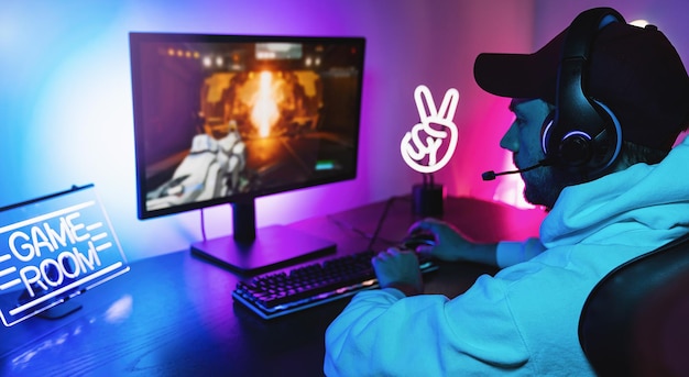 Gamer jogando videogame online de tiro em primeira pessoa em seu poderoso computador pessoal. Sala e PC têm luzes de néon coloridas. Jovem está jogando usando um boné em casa