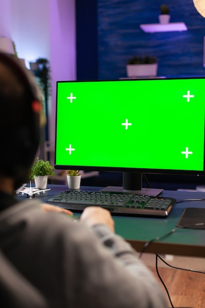 Gamer-Mann, der Videospiele auf dem Computer mit grünem Bildschirm spielt, der einen drahtlosen Joystick in einem professionellen Gaming-Studio hält. Spieler, der einen PC mit Mock-up isolierter Desktop-Streaming-Shooter-Spiele verwendet, die Headse tragen