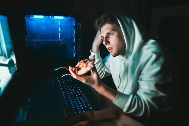 Gamer senta em casa à mesa do computador e come pizza