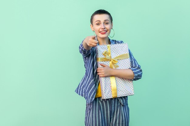 Ganador eres tú. Retrato de mujer joven hermosa feliz con pelo corto en traje a rayas de pie, abrazando el cuadro actual y señalando con el dedo a la cámara. Interior, aislado, foto de estudio, fondo verde