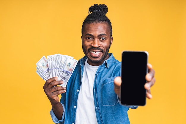 ¡Ganador! Joven rico feliz afroamericano negro en casual sosteniendo dinero billetes de un dólar con sorpresa aislado sobre fondo de pared amarilla. Usando teléfono móvil. Espacio de copia de pantalla.