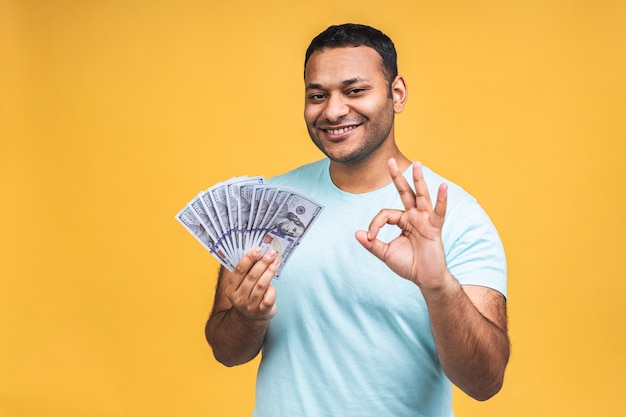 ¡Ganador! Joven rico feliz afroamericano negro indio en casual sosteniendo dinero billetes de un dólar con sorpresa aislado sobre fondo de pared amarilla.