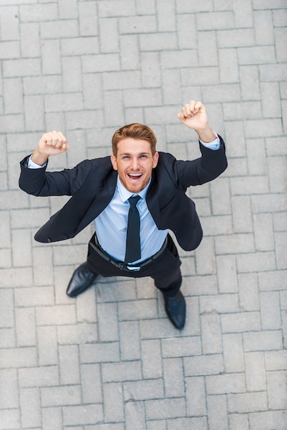 Ganador de todos los días. Vista superior del joven feliz en ropa formal manteniendo los brazos levantados y expresando positividad mientras está de pie al aire libre