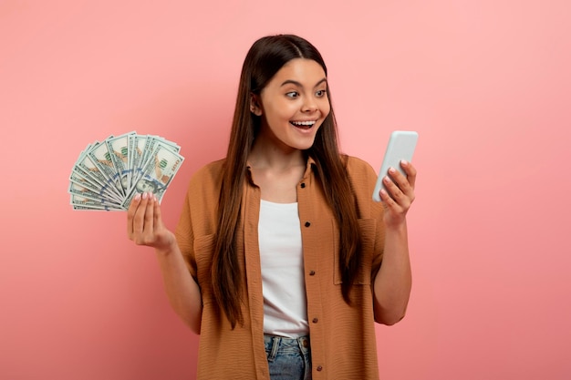 Foto ganar dinero en línea adolescente feliz sosteniendo dinero en efectivo y usando un teléfono inteligente