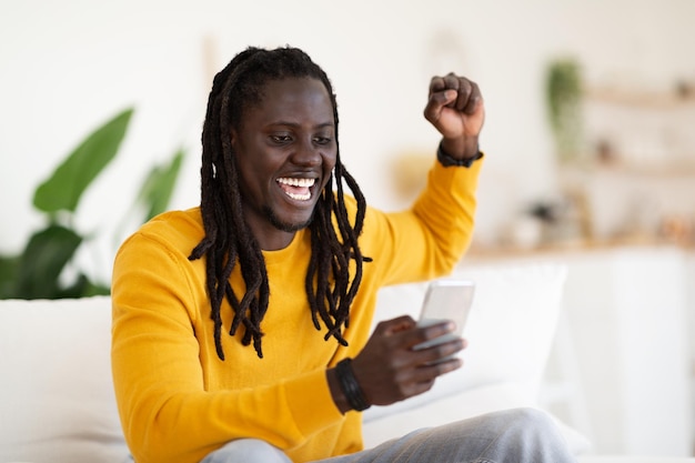 Ganar en línea Un hombre negro rebosante de alegría celebrando el éxito con un teléfono inteligente en casa