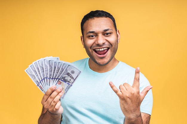 Ganhador! Jovem rico feliz afro-americano indiano negro casual segurando dinheiro notas de dólar com surpresa isolada sobre o fundo da parede amarela.