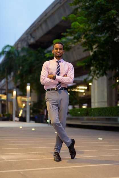 Ganzaufnahme des hübschen jungen afrikanischen Geschäftsmannes draußen in der Stadt nachts