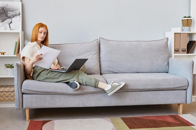 Ganzaufnahme einer erwachsenen rothaarigen Frau, die Laptop auf dem Sofa verwendet, während sie von zu Hause aus kopiert wird