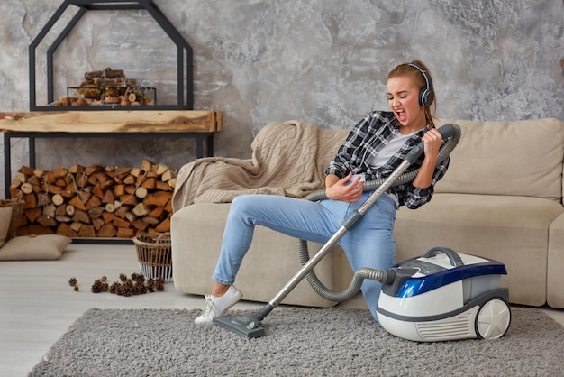 Ganzaufnahme einer fröhlichen jungen frau der 20er jahre, die über kopfhörer musik hört und spaß mit dem staubsauger im haus hat.