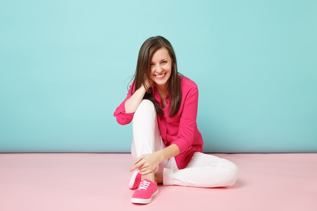 Ganzaufnahme einer lächelnden jungen Frau in rosa Hemdbluse, weiße Hose auf dem Boden sitzend