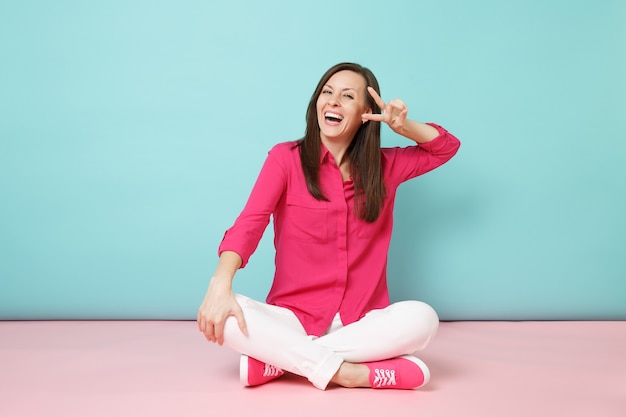 Ganzaufnahme einer lächelnden jungen Frau in rosa Hemdbluse, weiße Hose auf dem Boden sitzend