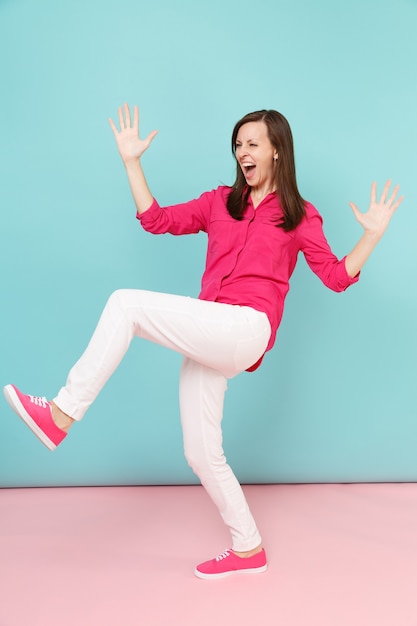 Ganzaufnahme einer wütenden, lustigen jungen hübschen Frau in rosa Hemdbluse, weiße Hose, die isoliert auf hellrosa blauer Pastellwand posiert.