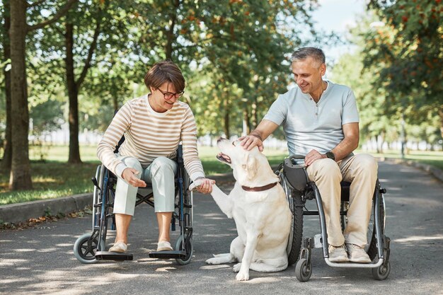 Ganzaufnahme eines erwachsenen Paares in Rollstühlen, das mit Hund im Park im Freien spielt, Platz kopieren