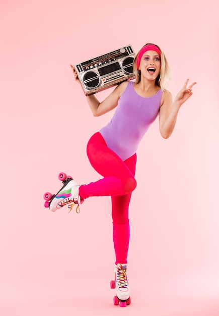 Ganzaufnahme eines fröhlichen jungen blonden Mädchens, das Retro-Sportkleidung und Rollschuhe trägt, die isoliert über rosa Wand posiert und Boombox hält