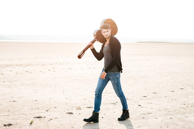 Ganzaufnahme eines jungen zufälligen Mannes, der am Strand spazieren geht und Gitarre auf seiner Schulter hält