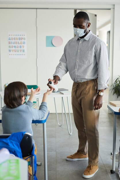 Ganzaufnahme eines männlichen Lehrers, der die Hände von Kindern im Schulklassenzimmer desinfiziert, Kovid-Sicherheitsmaßnahmen