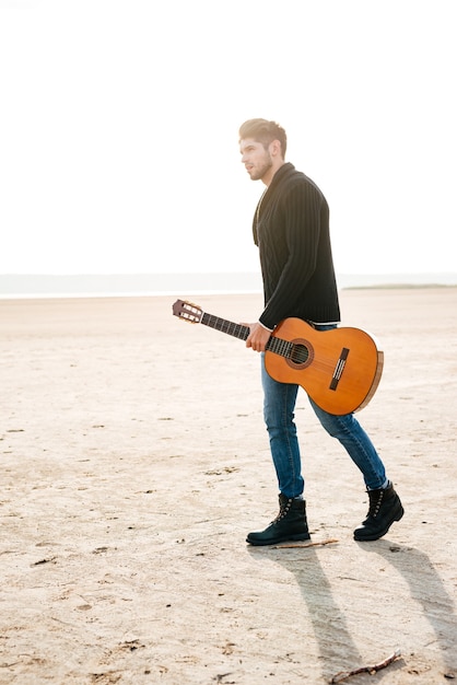 Ganzaufnahme eines männlichen Musikers, der mit Gitarre am Strand spazieren geht