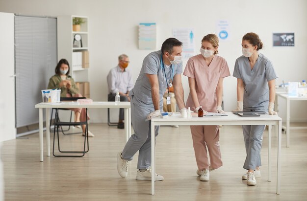 Ganzaufnahme von drei Ärzten, die Masken tragen, die in der medizinischen Klinik am Schreibtisch stehen, Kopierraum