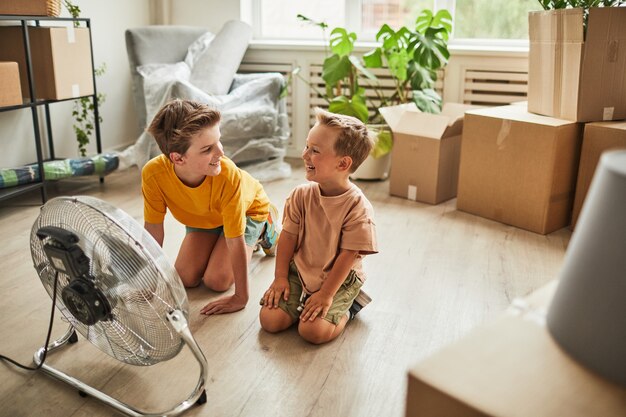 Ganzaufnahme von zwei Jungen, die am Ventilator sitzen und sich in der Hitze erfrischen, während die Familie in neue...