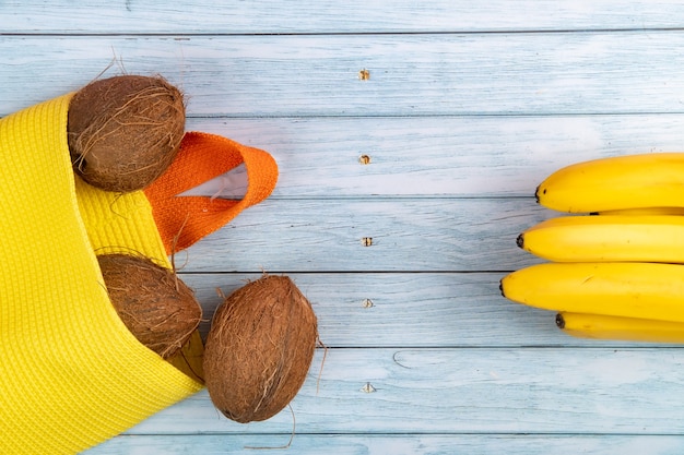 Ganze Kokosnüsse liegen in einem gelben Sack und Bananen auf blauem Holzhintergrund.