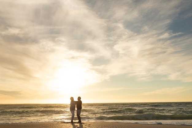 Ganze Länge eines gemischtrassigen Seniorenpaares, das bei Sonnenuntergang am Strand gegen den Himmel tanzt