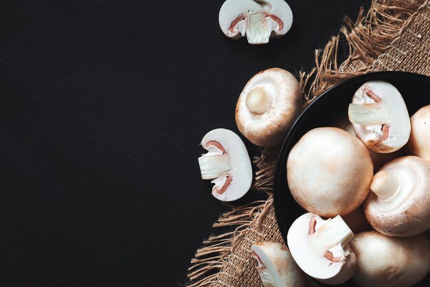 Ganze und geschnittene Champignons und Kräuter auf schwarzem Hintergrund. Nahaufnahme, flache Lage, Draufsicht. Lebensmittelhintergrund, selektiver Fokus, Kopienraum.