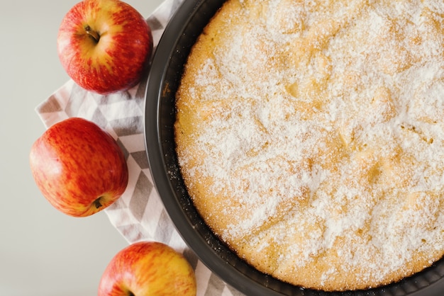 Ganzer Apfelkuchen mit Puderzucker auf einem Küchentuch serviert mit frischen roten Äpfeln