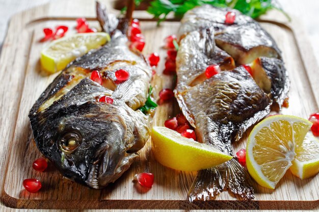 Ganzer gebackener Dorado-Fisch gefüllt mit frischer Petersilie auf einem Schneidebrett mit Zitronenspalten und Granatapfelkernen auf weißem Holztisch, Nahaufnahme, Draufsicht