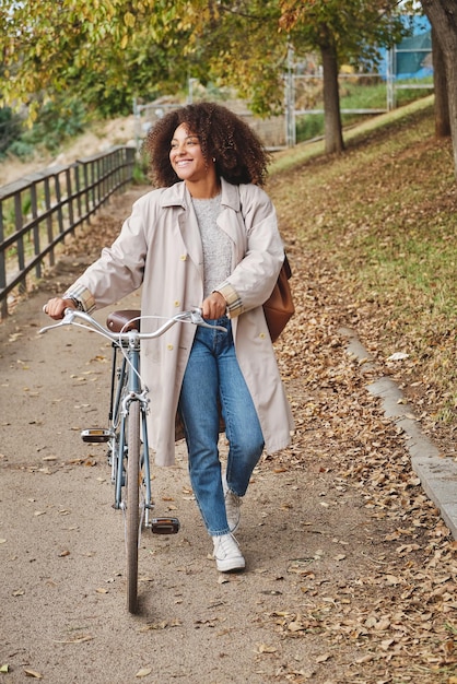 Ganzer Körper einer glücklichen jungen afroamerikanischen Frau mit Fahrrad, die beim Spaziergang im Herbstpark wegschaut