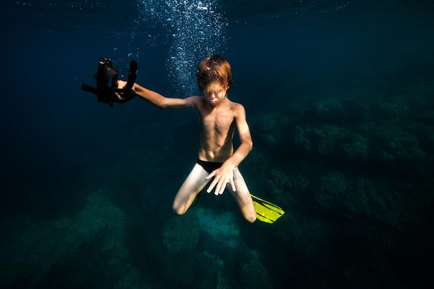 Ganzer Körper eines hemdlosen Teenagers mit Flossen und Schnorchelmaske in der Hand, der mit geschlossenen Augen unter Wasser taucht