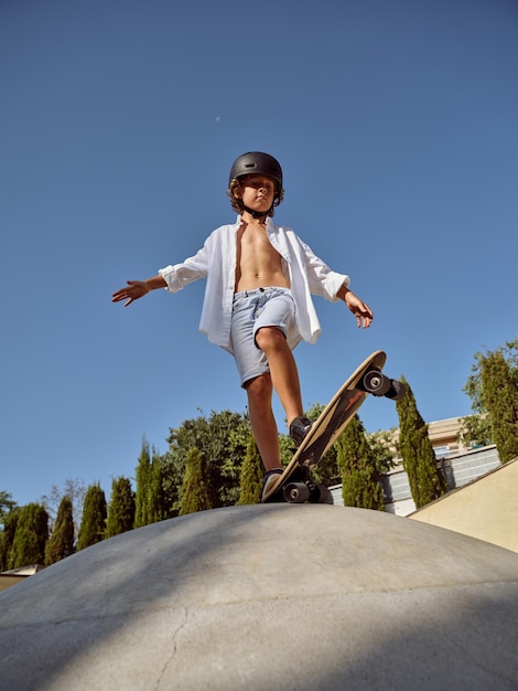 Ganzer Körper eines kleinen Jungen in lässigem Outfit und Helm, der in die Kamera blickt, während er auf einem Skateboard vor wolkenlosem blauen Himmel steht