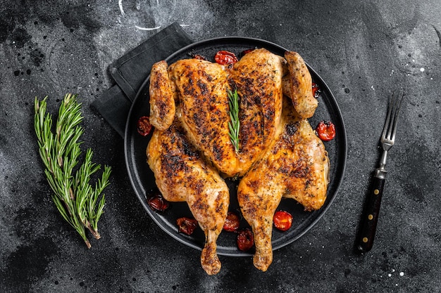 Ganzes Huhn des Tabaks auf Teller mit Kräutern und Tomate. Schwarzer Hintergrund. Ansicht von oben.