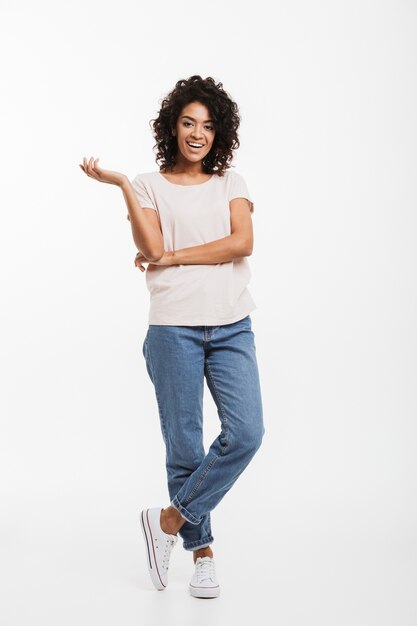 Foto ganzes porträt der lächelnden amerikanischen frau, die jeans und t-shirt trägt, die mit verschränkten armen stehen und hand beiseite gestikulieren, lokalisiert über weißer wand