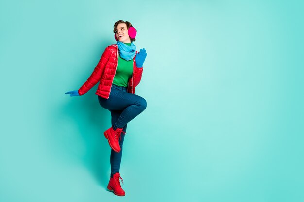 Ganzkörper-Profilfoto der lustigen Dame genießen sonnige Wintertagspaziergang Straße gute Laune tragen lässig roten Mantel Schal rosa Ohrhüllen Hosen Stiefel isoliert blaugrün Farbe Wand