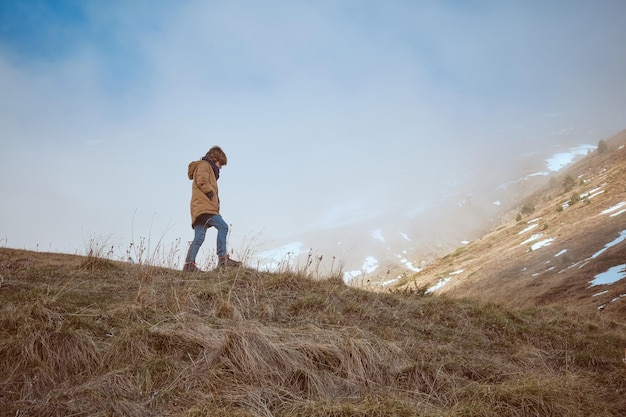 Ganzkörper-Seitenansicht eines Jungen in warmer Kleidung, der auf einer Wiese in der Nähe eines Berghangs mit Raureif in der Natur spaziert