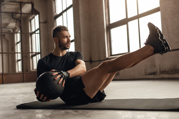Ganzkörper-Seitenansicht eines starken erwachsenen bärtigen Mannes in Sportbekleidung, der während des Bauchmuskeltrainings im Fitnessstudio sitzende Seitendrehübungen mit Medizinball macht