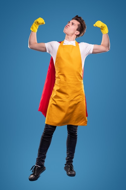 Ganzkörper selbstbewusster junger Mann in gelber Schürze und Handschuhen mit Superheldenmantel, der Stärke zeigt und ein lustiges Gesicht macht, während er den Reinigungsservice vor blauem Hintergrund darstellt